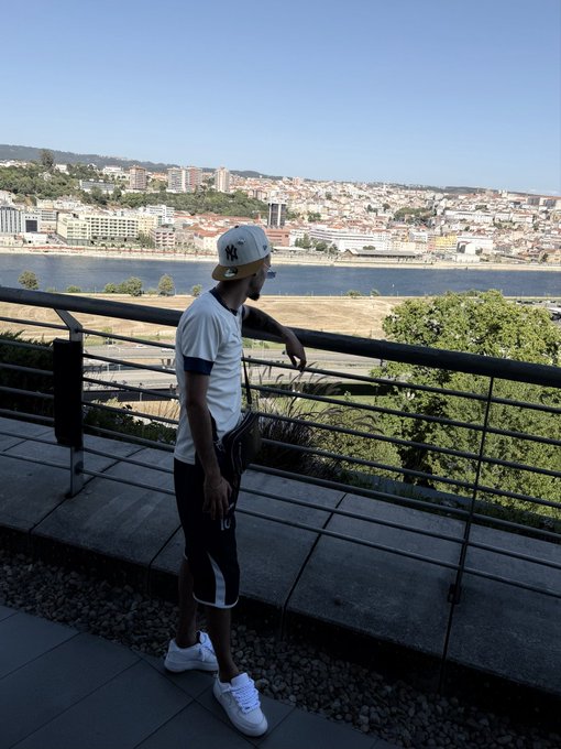 A man wearing a white t-shirt, black shorts, white sneakers, and a New York Yankees cap leans against a metal railing overlooking a river with a city skyline in the background.