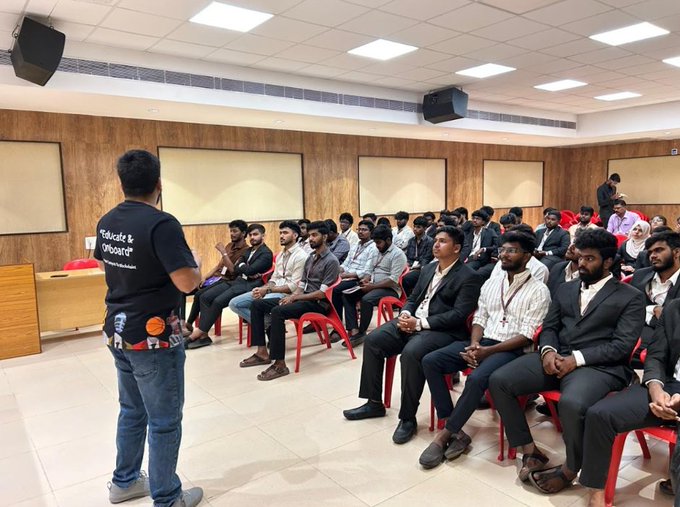 A classroom or seminar setting where a person in a black 'Educate & Onboard' t-shirt stands in front of an audience of seated attendees, mostly young men in business attire.