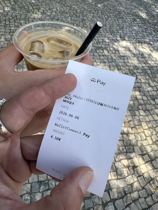 The image shows a person holding a receipt for their coffee purchase. The receipt has a phone number and a website listed on it, indicating that they have just paid with a mobile payment app or service. The person's hand is visible above the receipt, while the background features a cobblestone street and trees in the distance.
