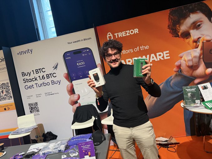 A man with curly hair and glasses stands at a Bitcoin/crypto event booth holding up a Trezor hardware wallet box, with banners behind him advertising Invity 'Buy 1 BTC Stack 1.6 BTC Get Turbo Buy' and Trezor hardware wallets.