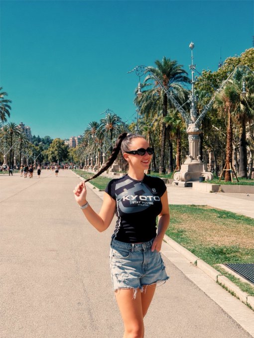 A smiling young woman wearing sunglasses and a black 'KYOTO' athletic shirt, playfully holding her braided hair while walking on a sunny palm-lined boulevard with ornate street lamps.