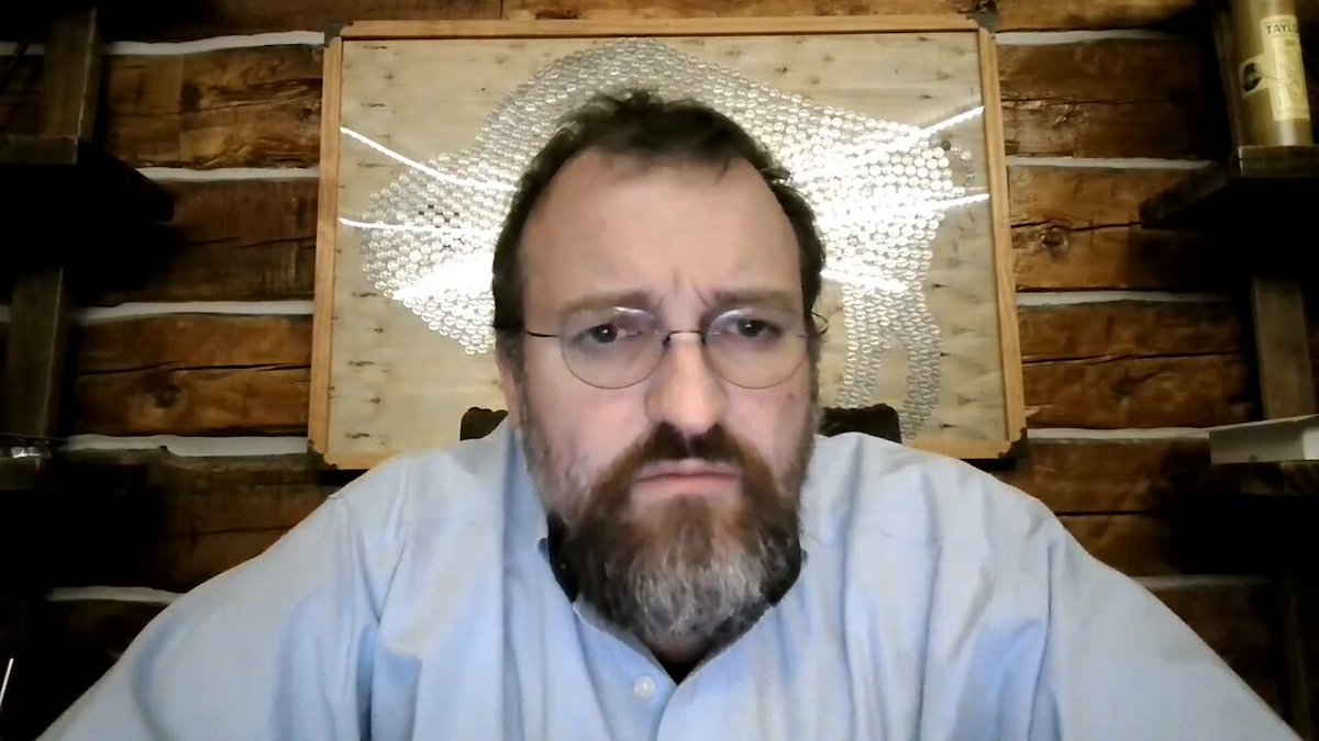 The image features a man wearing glasses sitting at a desk with his arms crossed over his chest. The background includes a wooden wall and several shelves filled with various items such as bottles and books. There are also some text elements, including "Cardano" written in the corner of the image and "we've grown in the last 6 months from about 11,000 users to 49,000 users".