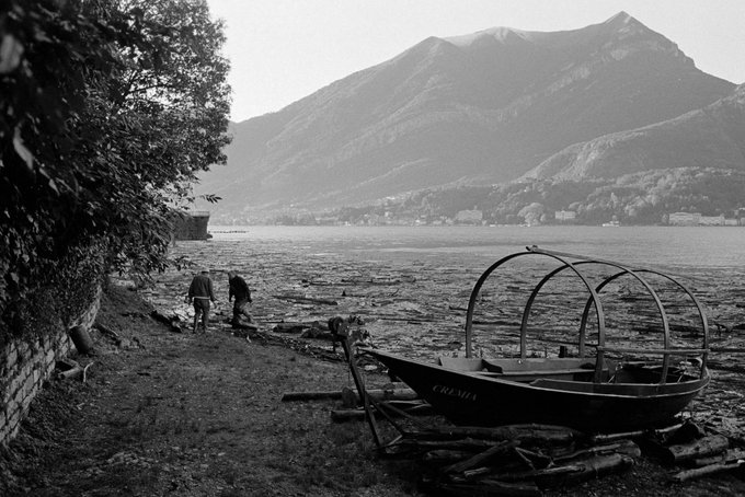 A black-and-white photograph shows two figures walking along a rocky shoreline with a beached boat named "CRESTA" in the foreground and misty mountains rising in the background, evoking a quiet Italian lakeside scene. The image reflects the photographer's diverse international portfolio—likely from the Italy series—offered as collectible works on ETH.