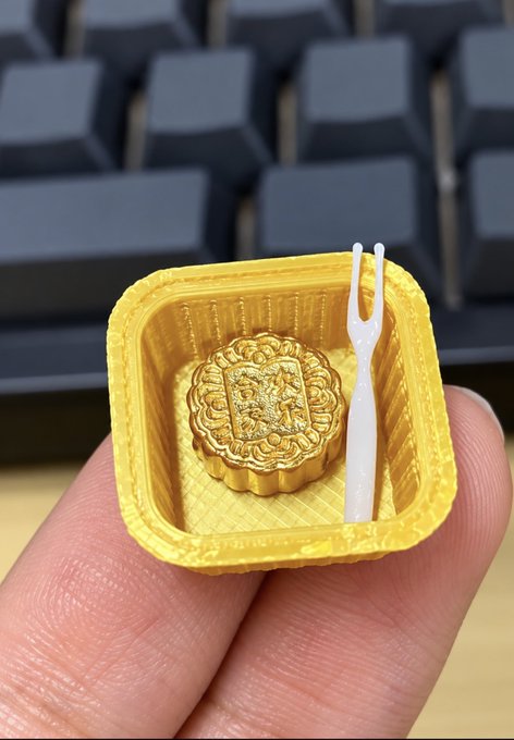 A tiny 3D-printed yellow container holds a miniature mooncake and a small white fork, resting on a person's fingertips.