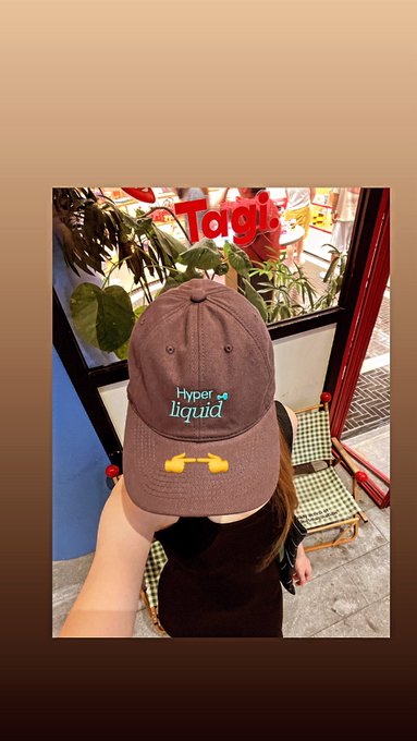 A hand holds up a brown 'hyper liquid' branded baseball cap with a yellow umbrella charm, displayed in front of a cafe window with a 'Tagu' sign.