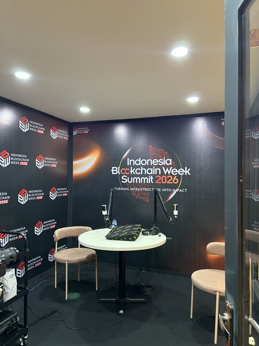 A live podcast setup at the Indonesia Blockchain Week Summit 2026, featuring a branded backdrop with the event's tagline "Turning Infrastructure Into Impact," a round table with microphones, and chairs ready for an upcoming discussion with builders, creators, and traders to be shared on Binance Square.