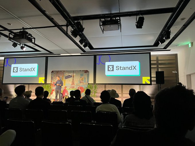 An indoor presentation or conference where a speaker stands on stage in front of a large screen displaying the 'StandX' logo, with audience members seated in the foreground watching.