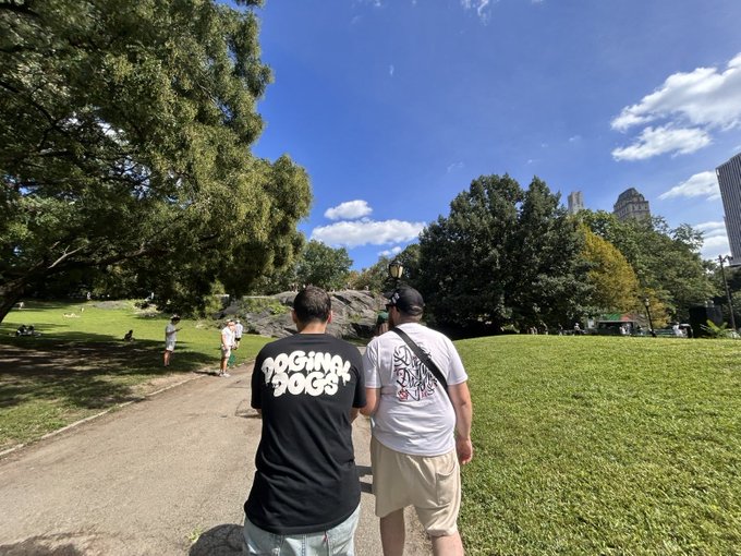 Two men walk along a sunny park path, one wearing a black "DOGMA DOGS" shirt and the other a white tee, both heading toward a bright blue sky as they enjoy a leisurely stroll in Central Park. The relaxed, optimistic "Gm Gm / Say it back??" vibe pairs perfectly with the fresh morning energy of the scene.