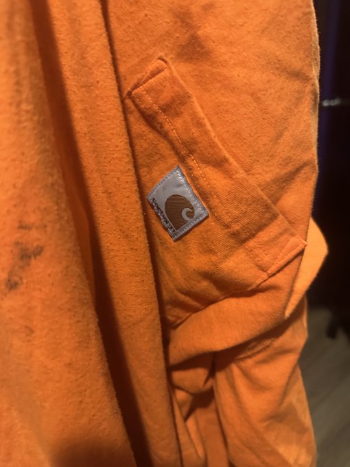 Close-up of an orange Carhartt jacket sleeve, focusing on the chest pocket with the brand's signature white logo patch.