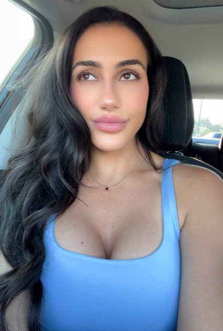 A woman with long dark wavy hair wearing a light blue tank top and a delicate necklace takes a selfie inside a car.