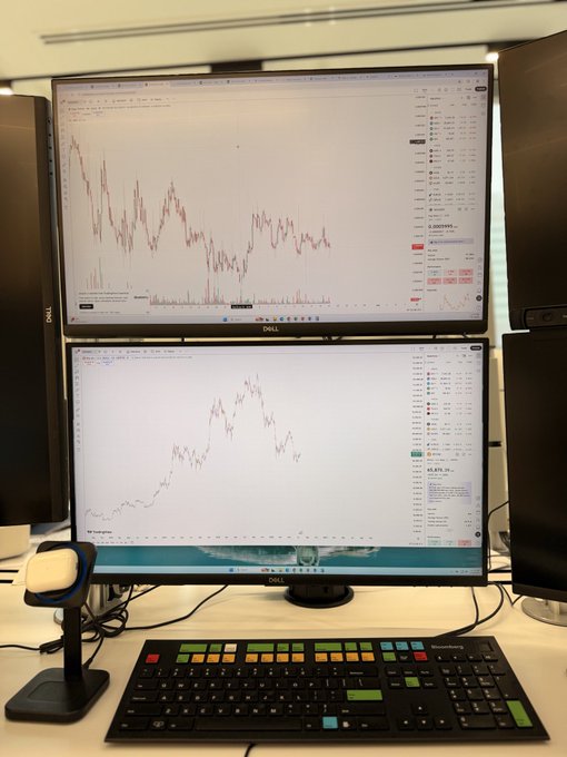 A dual-monitor trading workstation displaying cryptocurrency price charts (looks like BTC/USDT), accompanied by a specialized multi-colored hotkey keyboard and barcode scanner on a white desk.