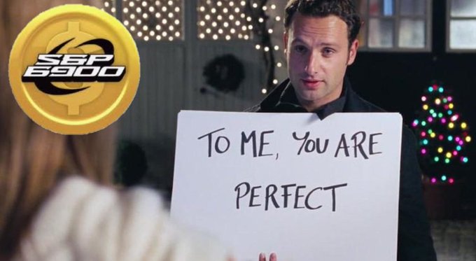 A scene from the movie 'Love Actually' showing a man holding a large white sign reading 'TO ME, YOU ARE PERFECT' in front of a woman, with a gold '$GBP 6900' coin overlaid in the corner and a Christmas tree in the background.