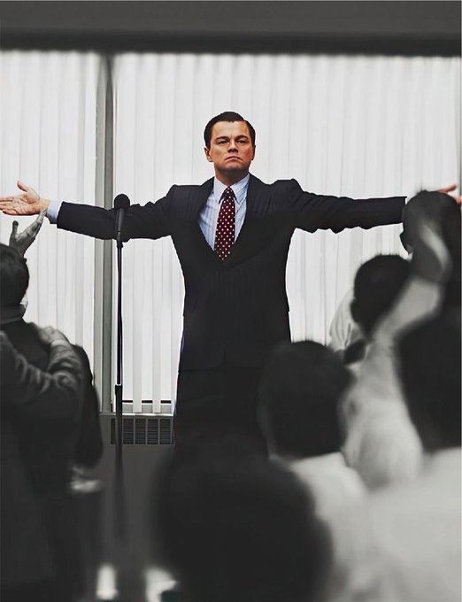 Leonardo DiCaprio in a suit stands with arms outstretched in front of an audience in what appears to be a scene from The Wolf of Wall Street.