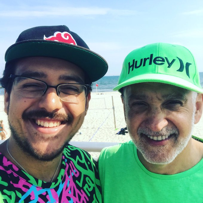 A selfie of two smiling men on a beach; the younger man wears a black cap and glasses, while the older man wears a bright green 'Hurley' cap and shirt.