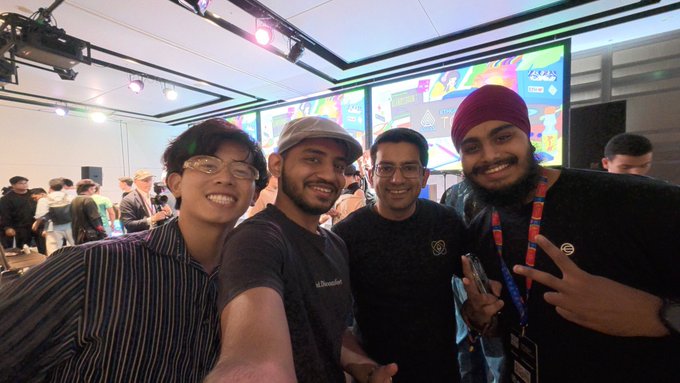 A group selfie of four young men smiling together at what appears to be an indoor event or convention with colorful displays in the background.