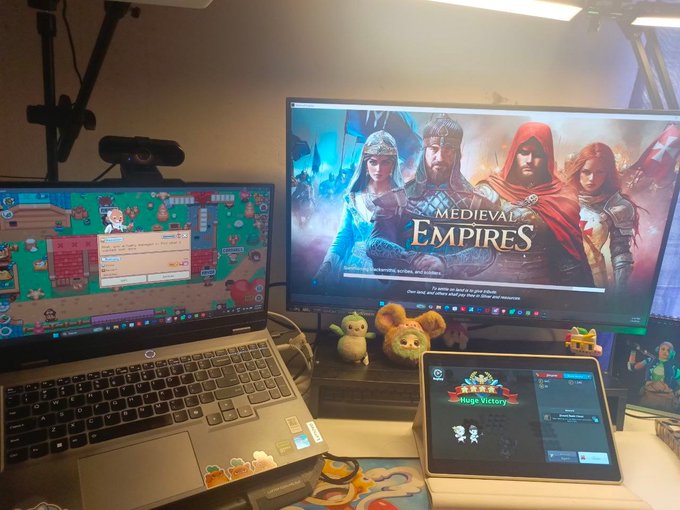 A desk setup showing a laptop and monitor with the game 'Medieval Empires' displayed, plus a tablet showing 'Huggy Wuggy' and various small plush toys and figurines.