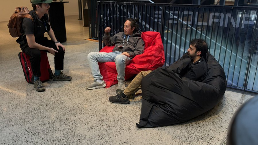 Three men relax in casual beanbag chairs in an indoor space near a railing with the text "REPUBLICAN" partially visible; one man wears a green cap and backpack, another sits in a red beanbag, and the third is wrapped in a black sleeping bag.