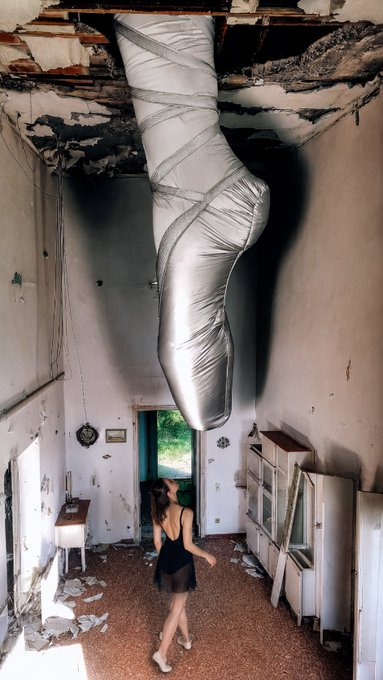 A surreal art installation showing a massive silver leg sculpture descending through a hole in the ceiling of a dilapidated building, with a person standing below.