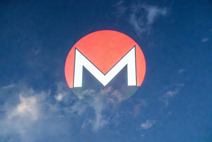 The Monero cryptocurrency logo—a stylized white 'M' inside a red circle—shown against a backdrop of blue sky with wispy white clouds.