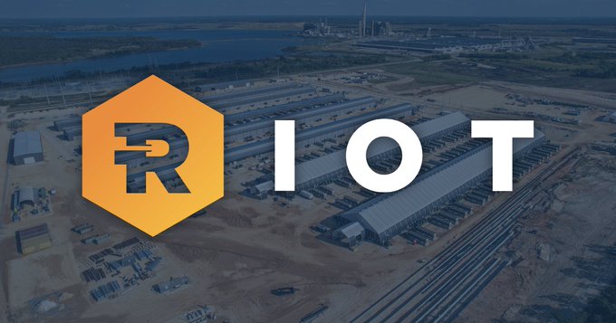 The image shows the Riot Platforms logo alongside the ticker "RIOT" overlaid on an aerial view of a large industrial facility, visually reinforcing the article about Riot's $9.1 billion contract with Anthropic to provide 191 MW of Texas data center capacity for AI over 20 years.