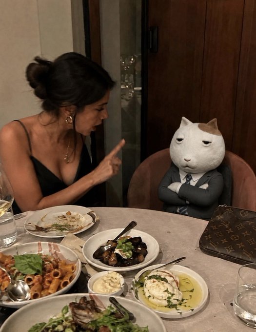 A woman at a restaurant table pointing her finger at a person wearing a white cat mask and a suit sitting across from her, surrounded by plates of food.