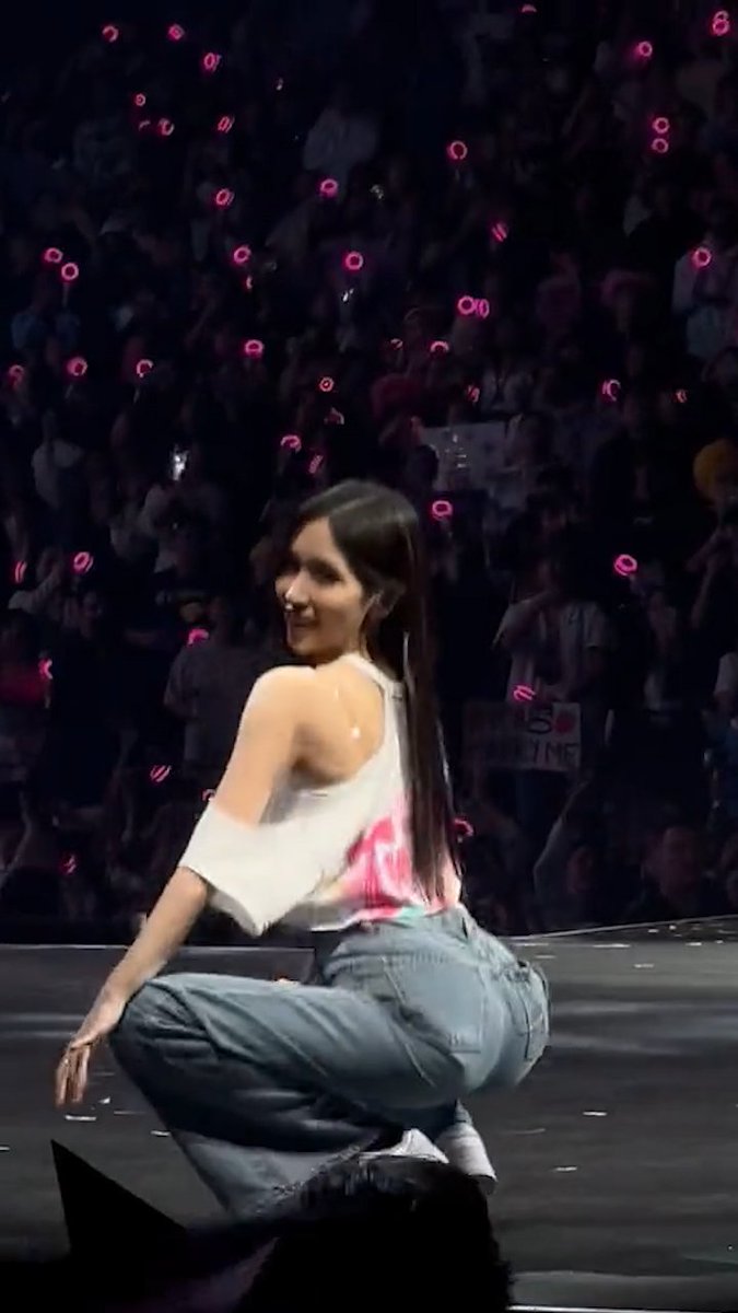 A performer with long dark hair, wearing a white off-shoulder top and light blue jeans, crouches on stage while turning to look over her shoulder with a smile. Behind her, a large crowd waves pink light sticks and holds a partially visible sign, connecting to the post about #MINA of #TWICE at a concert.