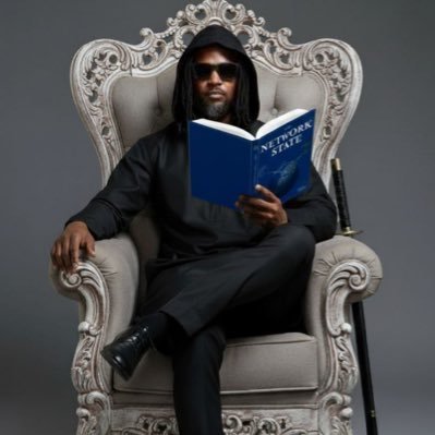 A man dressed in black with a hood, sunglasses, and dreadlocks sits regally on an ornate white throne while reading a blue book titled "NETWORK STATE," accompanied by a sword at his side—powerfully symbolizing the "Chairman" role and visionary leadership celebrated in the birthday tribute.