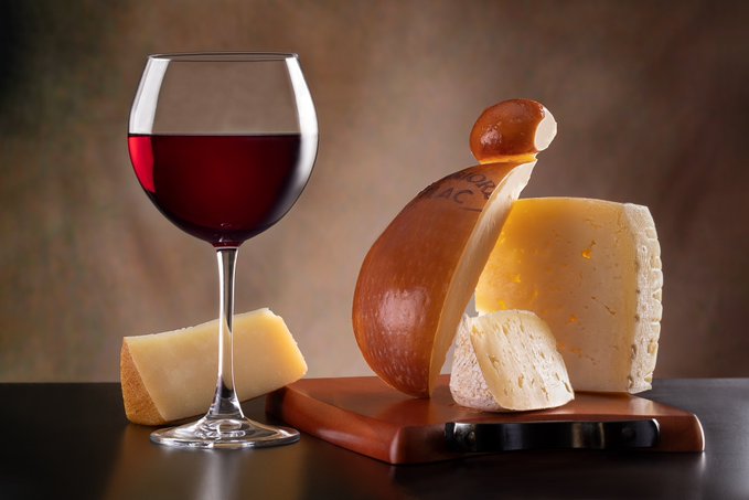 A still life arrangement featuring a glass of red wine alongside various cheeses and a knife on a wooden cutting board.