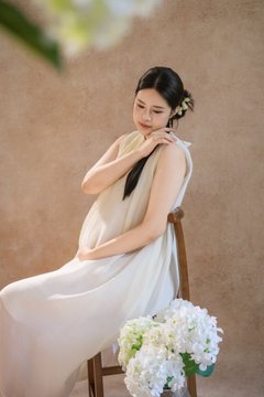 A pregnant woman with dark hair styled in a braid sits on a wooden chair wearing a flowing white dress, holding white hydrangeas.