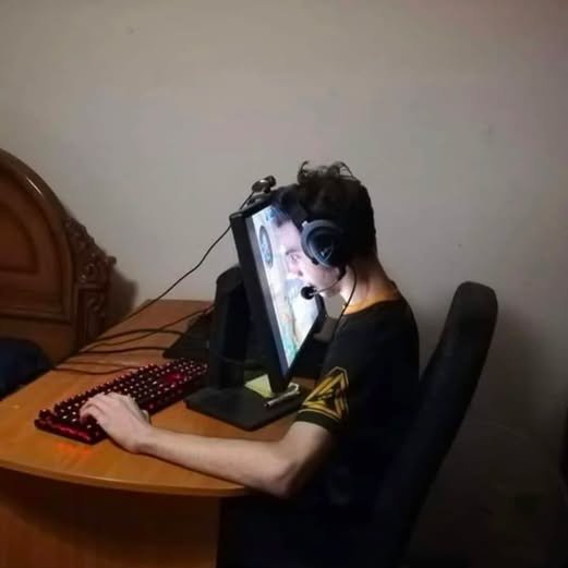 A young person wearing headphones sits at a wooden desk using a red backlit keyboard and monitor, appearing focused on gaming.