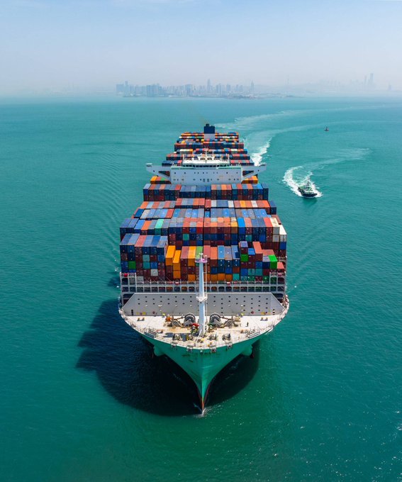 A massive container ship loaded with multicolored cargo containers sails through turquoise waters toward a distant city skyline, accompanied by a small escort vessel. The image visually reinforces the post's message about maritime logistics—a backbone of global trade and a compelling candidate for blockchain-based real-world asset tokenization.