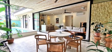 An open-plan living and dining area with a pool visible through large glass sliding doors, featuring modern furniture and a round dining table with wooden chairs.