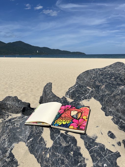 An open coloring book with a colorful bird and floral illustration resting on dark rocks at a sandy beach, with the ocean and mountains in the background under a blue sky.