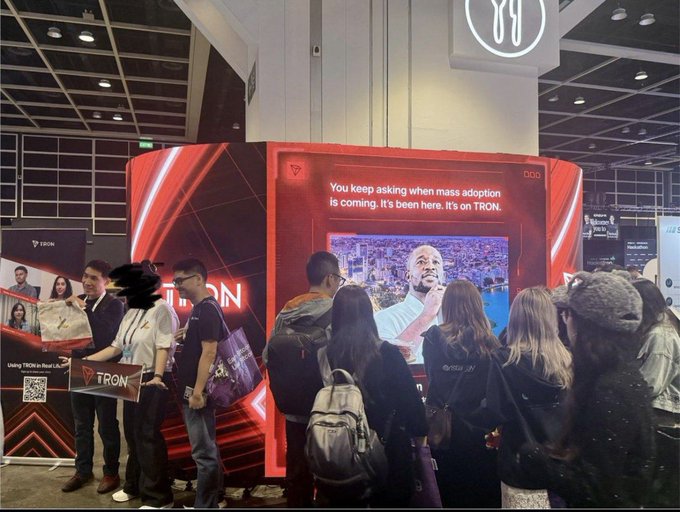 In this image, a group of people are gathered around an electronic display at a convention or exhibition area. There are several individuals standing close together with one person holding up their phone, possibly taking a picture of something interesting happening on the screen. The electronic device in question appears to be playing some videos, as there is text displayed above it that says "You keep asking when most adaptation is coming. It's been here, on an TRON Không Chỉ Mở Rộng Hệ Sinh Thái". The group seems engaged and interested in the content being presented, creating a lively atmosphere at the event.