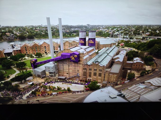 An aerial view of a large industrial building decorated with purple banners featuring a yellow 'A' logo, with crowds of people gathered below.
