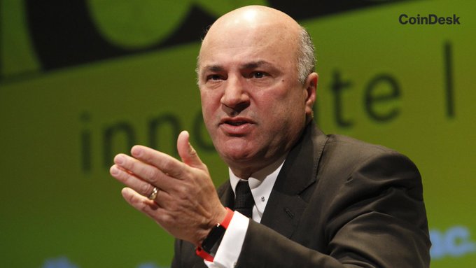 A bald man in a dark suit speaking at what appears to be a CoinDesk conference, with a green background featuring partial text.