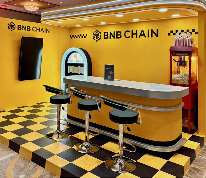 A vibrant BNB Chain-themed diner booth with yellow walls, checkered flooring, and a retro bar counter, where the prompt "What are you ordering?" invites attendees to choose from the crypto-themed setup, complete with a popcorn machine and bar stools.

The image relates to the post by playfully framing a crypto/blockchain event booth as a classic American diner, encouraging engagement by asking what attendees would "order" from the BNB Chain menu.