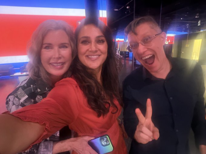 Three people pose for a cheerful selfie in what appears to be a modern studio space, with the woman in the center wearing a rust-colored dress and holding a smartphone, flanked by a blonde woman on the left and a bespectacled man on the right flashing a peace sign with an enthusiastic expression. The vibrant setting aligns with the post's description of visiting the "extraordinarily exciting" Beeple (Mike Winkelmann) studio for a memorable first-time visit.