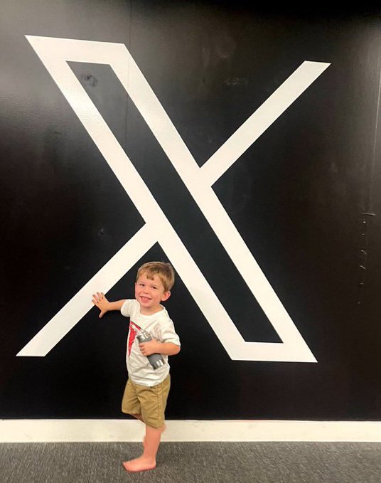 A young barefoot boy in a white shirt and khaki shorts smiles while posing in front of a large white X logo on a black wall.