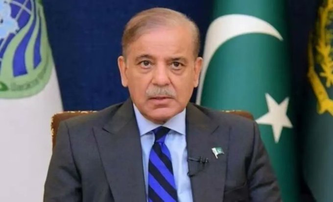 A man in a gray suit and blue striped tie speaks at a podium with Pakistani flags behind him.