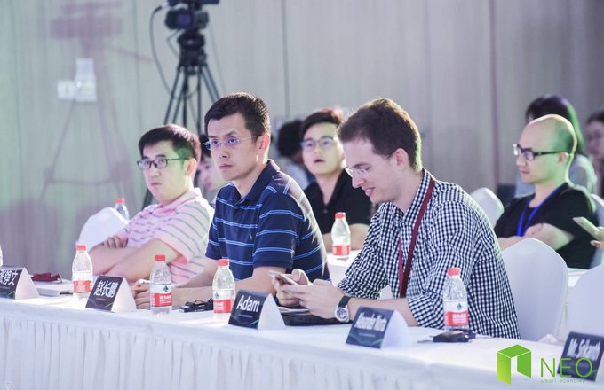 Several men seated at a conference table with name placards (including 'Adam' and 'Adam Wei') at a NEO event, with water bottles and broadcasting equipment visible.