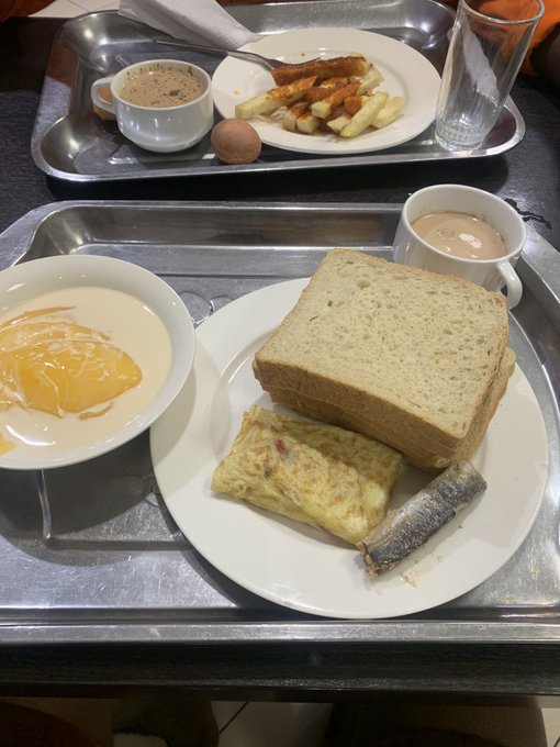 Two metal cafeteria trays holding breakfast items including toast, an omelet, a small fish, a bowl of yogurt or dessert, coffee, and a boiled egg.