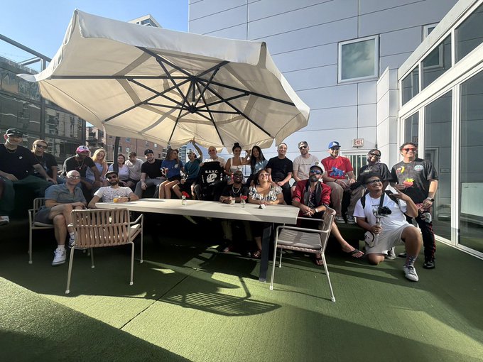 A diverse group of people gathers outdoors under a large beige umbrella on a rooftop patio with artificial green turf, posing for a group photo around a white table.