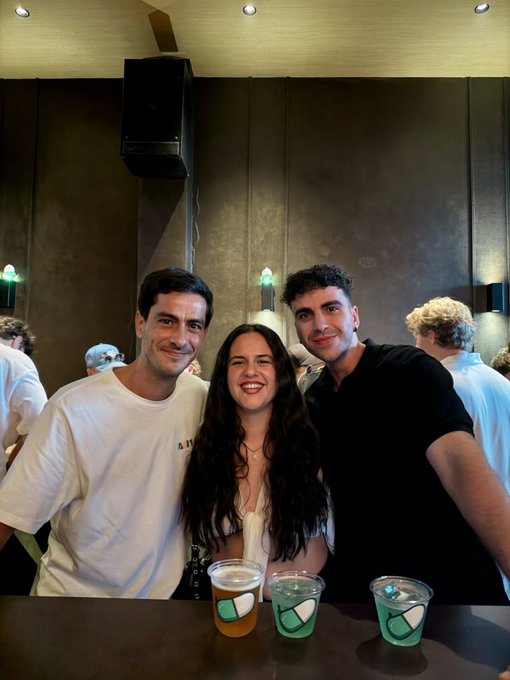Three young people—two men and a woman—smile for a photo at a bar or event, with three plastic cups of drinks lined up on the counter in front of them.