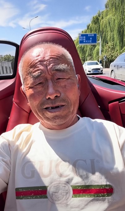 A close-up selfie of an elderly man with a mustache, eyes closed, wearing a white Gucci sweatshirt, sitting in a red leather car seat with a street scene visible in the background.