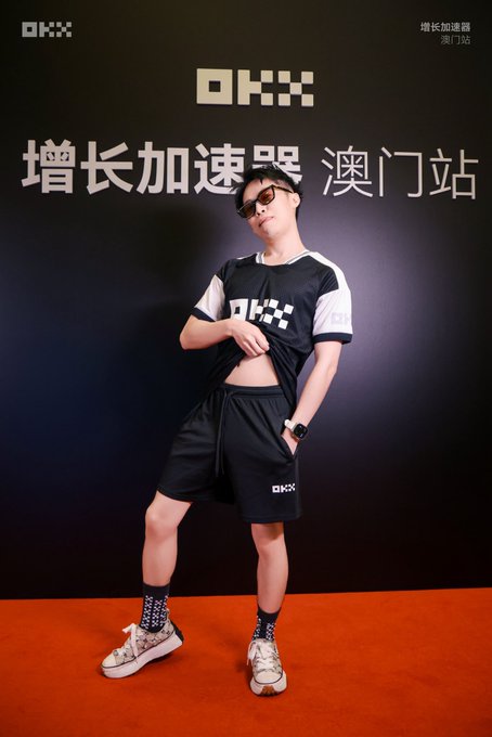 The image shows a person posing on an orange carpet in front of a black backdrop featuring "OKX" branding and Chinese text "增长加速器 澳门站" (Growth Accelerator Macau Station), wearing a black and white OKX-branded outfit. This relates to the caption about attending the OKX Macau Growth Station event and the red carpet mentioned at the banquet.