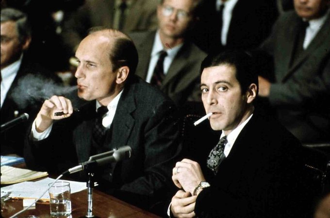 A vintage photograph of two men in dark suits sitting at a formal hearing table with microphones; one has a cigar, the other is smoking a cigarette, with other suited figures visible in the background.