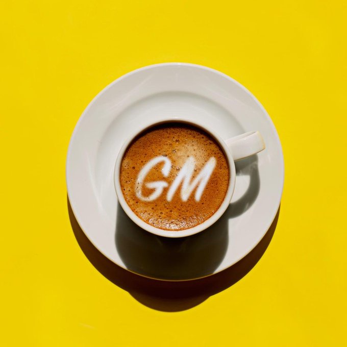 A top-down view of a white coffee cup on a saucer against a bright yellow background, with 'GM' written in foam on the coffee surface.