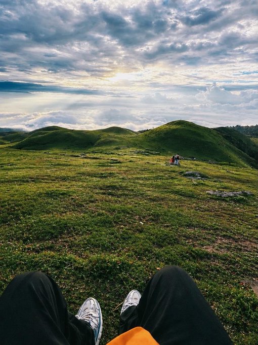 A first-person perspective photo taken from a grassy hilltop, showing the photographer's legs and sneakers in the foreground, looking out over rolling green hills under a dramatic, cloudy sky with the sun breaking through.