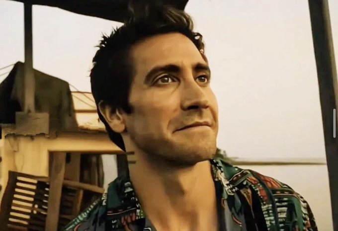 A close-up of a man with dark wavy hair wearing a colorful patterned shirt, looking off to the side with a slight smile, set against an outdoor background.